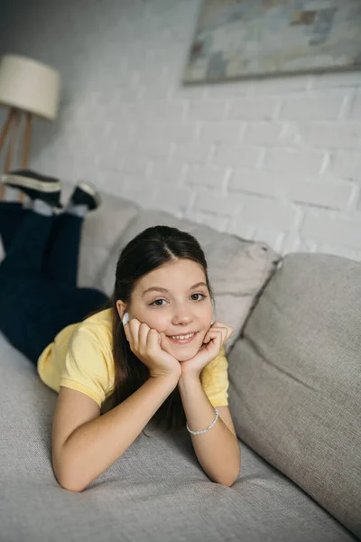 Menina alegre ouvir música no fone de ouvido sem fio enquanto deitado no sofá com as mãos perto do rosto — Fotografia de Stock