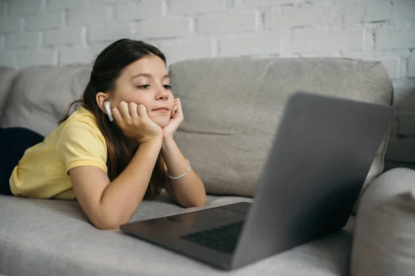 Брюнетка дівчина в бездротових навушниках дивиться на розмитий ноутбук, лежачи на затишному дивані вдома — Stock Photo