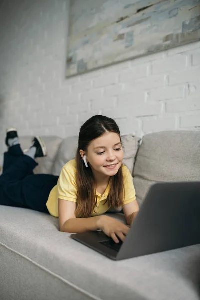 Smiling girl in wireless earphone lying on soft couch and typing on laptop — Stock Photo