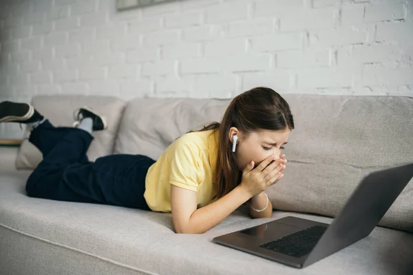 Bored girl in wireless earphone yawning near laptop on comfortable couch in living room — Stock Photo