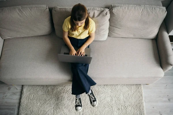 Vista superior de la niña preadolescente utilizando el ordenador portátil en el sofá moderno en casa - foto de stock