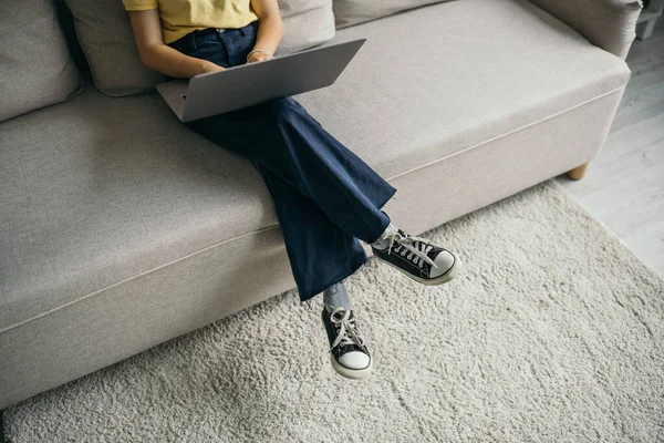 Visão de alto ângulo da criança cortada com laptop sentado no sofá confortável em casa — Fotografia de Stock