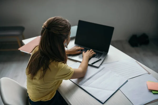 Visão de alto ângulo da menina na digitação de fone de ouvido no laptop com tela em branco perto de caderno e papéis — Fotografia de Stock