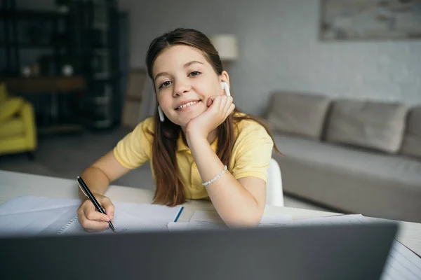 Fröhliches Mädchen mit drahtlosen Kopfhörern, Stift in der Hand und lächelnd in die Kamera auf verschwommenem Vordergrund — Stockfoto
