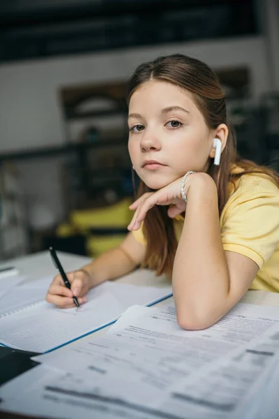 Дівчина в бездротових навушниках тримає ручку і дивиться на камеру, роблячи домашнє завдання — стокове фото