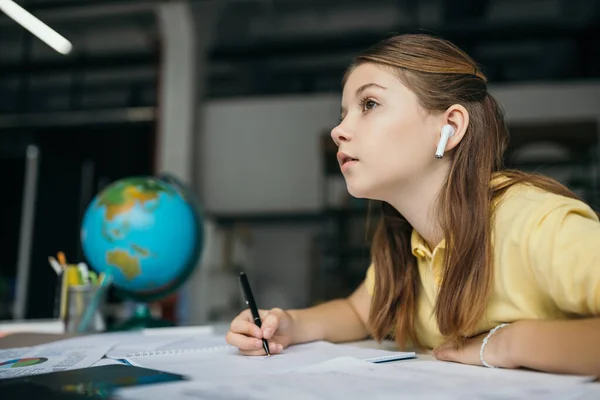 Side view of thoughtful girl in wireless earphone holding pen and looking away while doing homework — Stock Photo