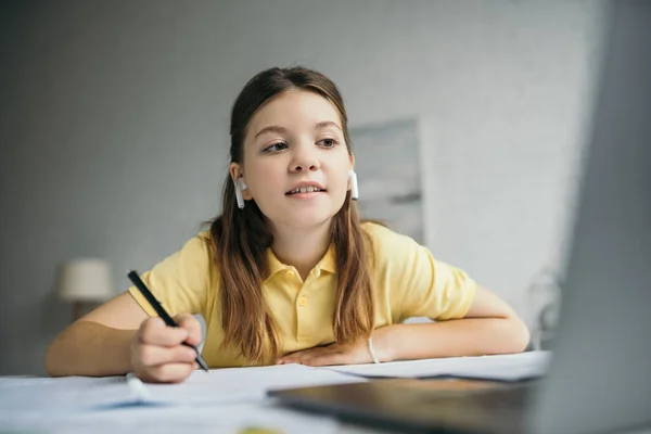 Усміхнена дівчина в бездротових навушниках тримає ручку і дивиться на ноутбук на розмитому передньому плані — стокове фото