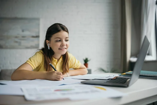 Усміхнена дівчина в бездротових навушниках тримає ручку і дивиться на ноутбук вдома — стокове фото