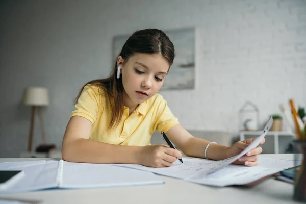 Brunette schoolgirl in wireless earphone writing and looking at copybook at home — Stock Photo