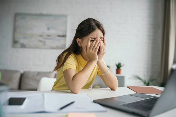 Exhausted kid with closed eyes covering face with hands while sitting near blurred laptop at home — Stock Photo