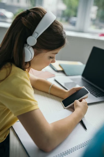Дівчина в навушниках тримає смартфон з порожнім екраном біля розмитого ноутбука — стокове фото