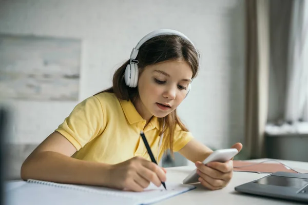 Mädchen mit Kopfhörer hält Smartphone in der Hand und schreibt in der Nähe eines verschwommenen Laptops — Stockfoto