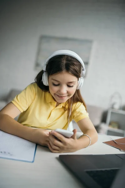Chica feliz en los auriculares que sostienen el teléfono móvil cerca de la computadora portátil borrosa mientras se aprende en casa - foto de stock