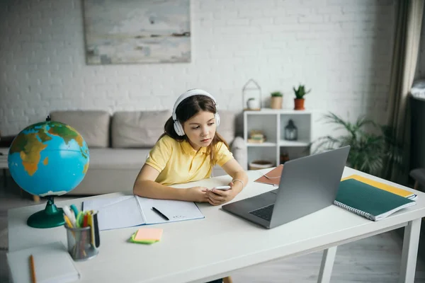 Preteen girl in headphones holding smartphone and looking at laptop near globe on table in living room — Stock Photo