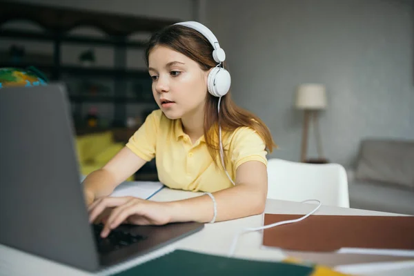 Brunette schoolgirl in wired earphones learning at home and typing on laptop — Stock Photo