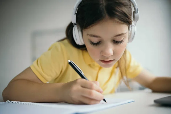 Chica enfocada en la escritura de auriculares con pluma durante la educación en casa - foto de stock