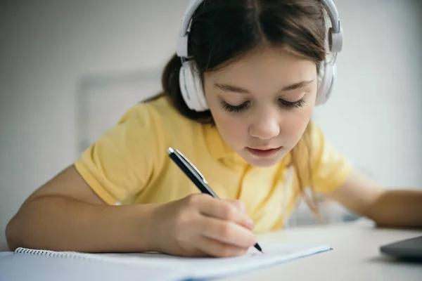 Ragazza concentrata in cuffie scrivere con penna durante la scuola a casa — Foto stock