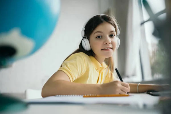 Menina pré-adolescente feliz em fones de ouvido fazendo lição de casa e sorrindo para a câmera em primeiro plano borrado — Fotografia de Stock