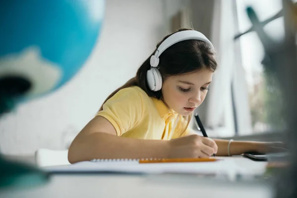 Schülerin mit Kopfhörer schreibt mit Stift, während sie zu Hause auf verschwommenem Vordergrund lernt — Stockfoto
