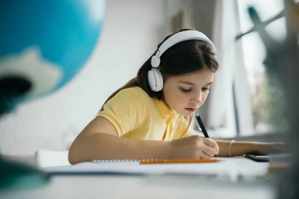 Schoolgirl in headphones writing with pen while learning at home on blurred foreground — Stock Photo