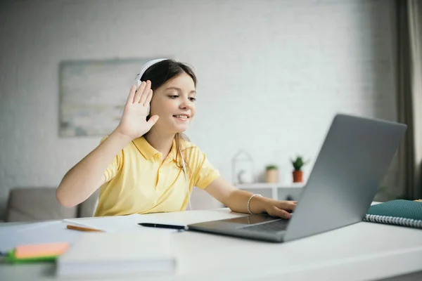 Улыбающаяся девушка в наушниках машет рукой возле ноутбука во время онлайн урока дома — стоковое фото