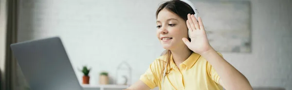 Preteen fille dans casque agitant la main et souriant pendant l'appel vidéo sur ordinateur portable, bannière — Photo de stock