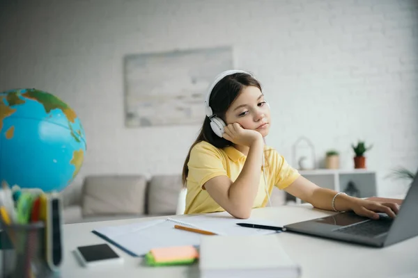 Pensive girl in headphones sitting near blurred laptop and globe while studying at home — Stock Photo