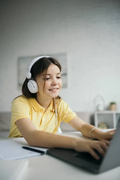 Cheerful schoolgirl in headphones studying at home and typing on laptop — Stock Photo