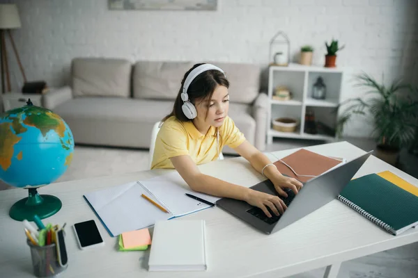 High angle view of girl in headphones typing on laptop near globe and copybooks — Stock Photo