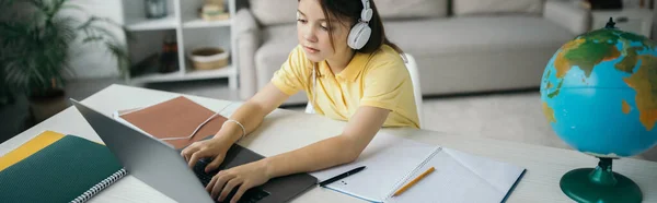 Высокий угол обзора девушки в наушниках, печатающих на ноутбуке рядом с ноутбуками и глобусом, баннер — стоковое фото