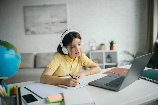 Fille dans les écouteurs regardant l'ordinateur et l'écriture dans le copybook pendant l'enseignement à domicile — Photo de stock
