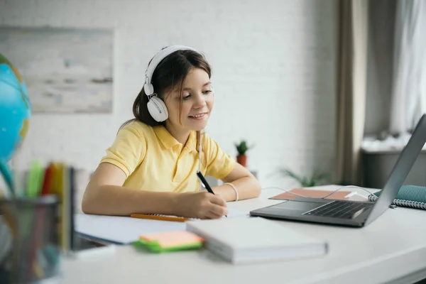 Souriant écolière écrit dans un cahier tout en étant assis dans des écouteurs près d'un ordinateur portable à la maison — Photo de stock