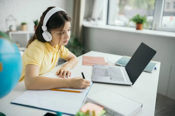 Child in headphones looking at laptop and writing in notebook at home — Stock Photo