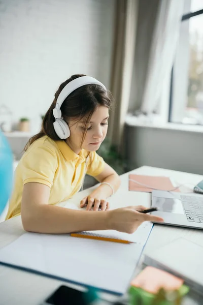 Colegiala en auriculares apuntando con el dedo mientras estudia cerca borrosa portátil en casa - foto de stock