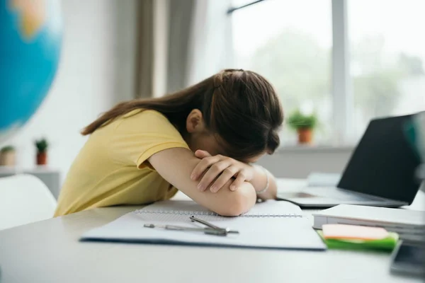 Side view of exhausted girl sleeping on copybook near laptop with blank screen on blurred background — Stock Photo
