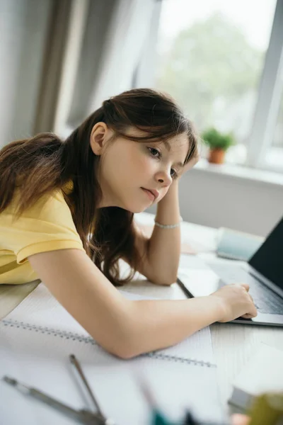 Nachdenkliches und gelangweiltes Mädchen sitzt neben Notizbuch und verschwommenem Laptop zu Hause — Stockfoto