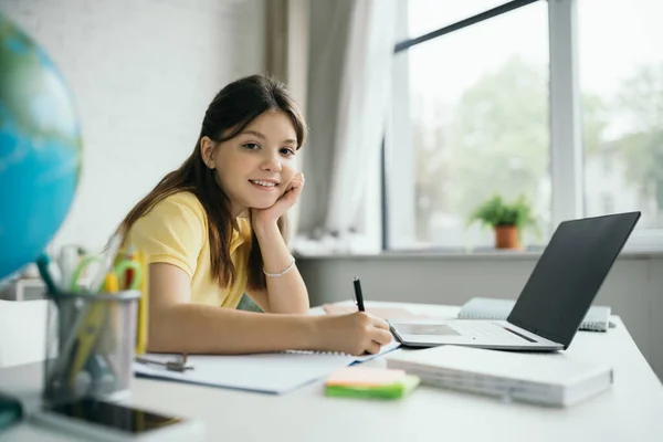 Cheerful girl holding pen and looking at camera near laptop with blank screen at home — Stock Photo