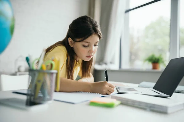 Brunette girl writing in notebook near laptop with blank screen at home — Stock Photo