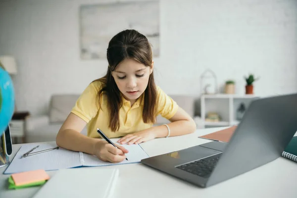 Preteen girl writing in notebook while learning near laptop in living room — Stock Photo