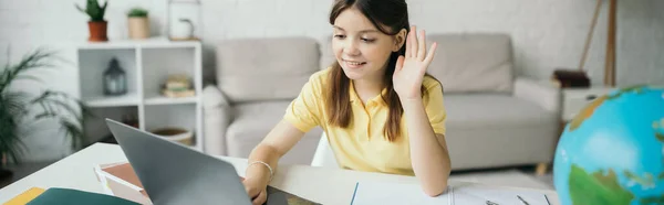 Alegre chica mirando a la computadora portátil y agitando la mano durante la lección en línea en casa, pancarta - foto de stock