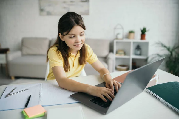 Sorridente ragazza bruna digitando sul computer portatile vicino notebook sul tavolo a casa — Foto stock