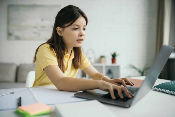 Preadolescente chica escribiendo en el ordenador portátil cerca borrosa copybooks durante la educación en casa - foto de stock