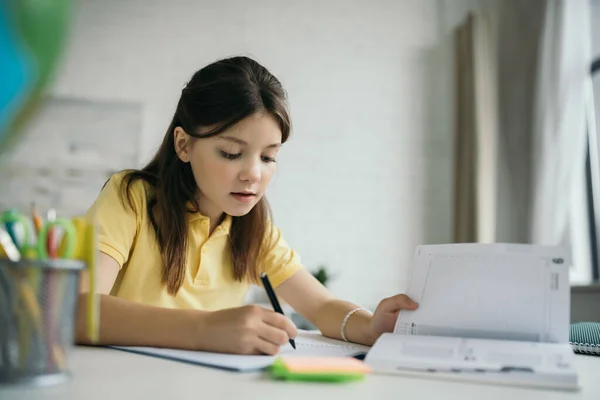 Preteen girl writing in notebook near diary on blurred foreground — Stock Photo