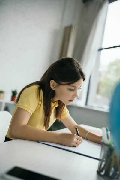 Konzentriertes Kind schreibt während der Hausaufgabenbetreuung in Notizbuch auf verschwommenem Vordergrund — Stockfoto