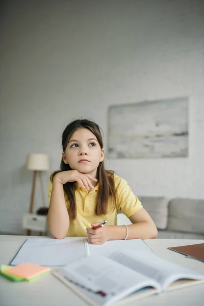 Menina pensativa segurando a mão perto do queixo e olhando para longe ao fazer lição de casa — Fotografia de Stock