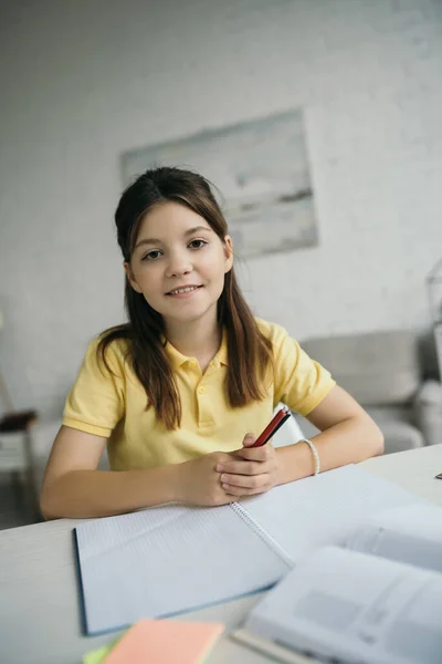 Веселая девушка держит ручки и смотрит в камеру рядом с пустым блокнотом дома — стоковое фото