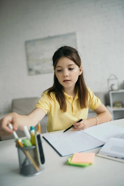 Брюнетка девушка выбирает ручку рядом с пустым блокнотом на столе в гостиной — стоковое фото