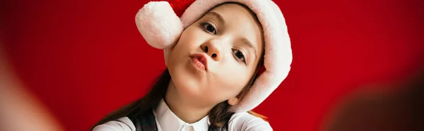 Funny child in santa hat looking at camera and pouting lips isolated on red, banner — Stock Photo