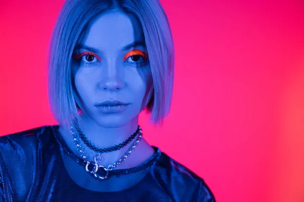 Portrait of trendy woman with necklaces and fluorescent makeup in blue neon light on deep pink background — Stock Photo