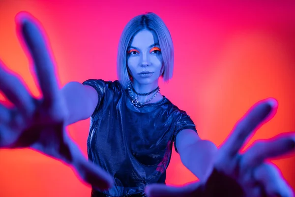 Stylische Frau mit ausgestreckten Händen, die in blauem Neonlicht auf korallenfarbenem und rosa Hintergrund in die Kamera blickt — Stockfoto